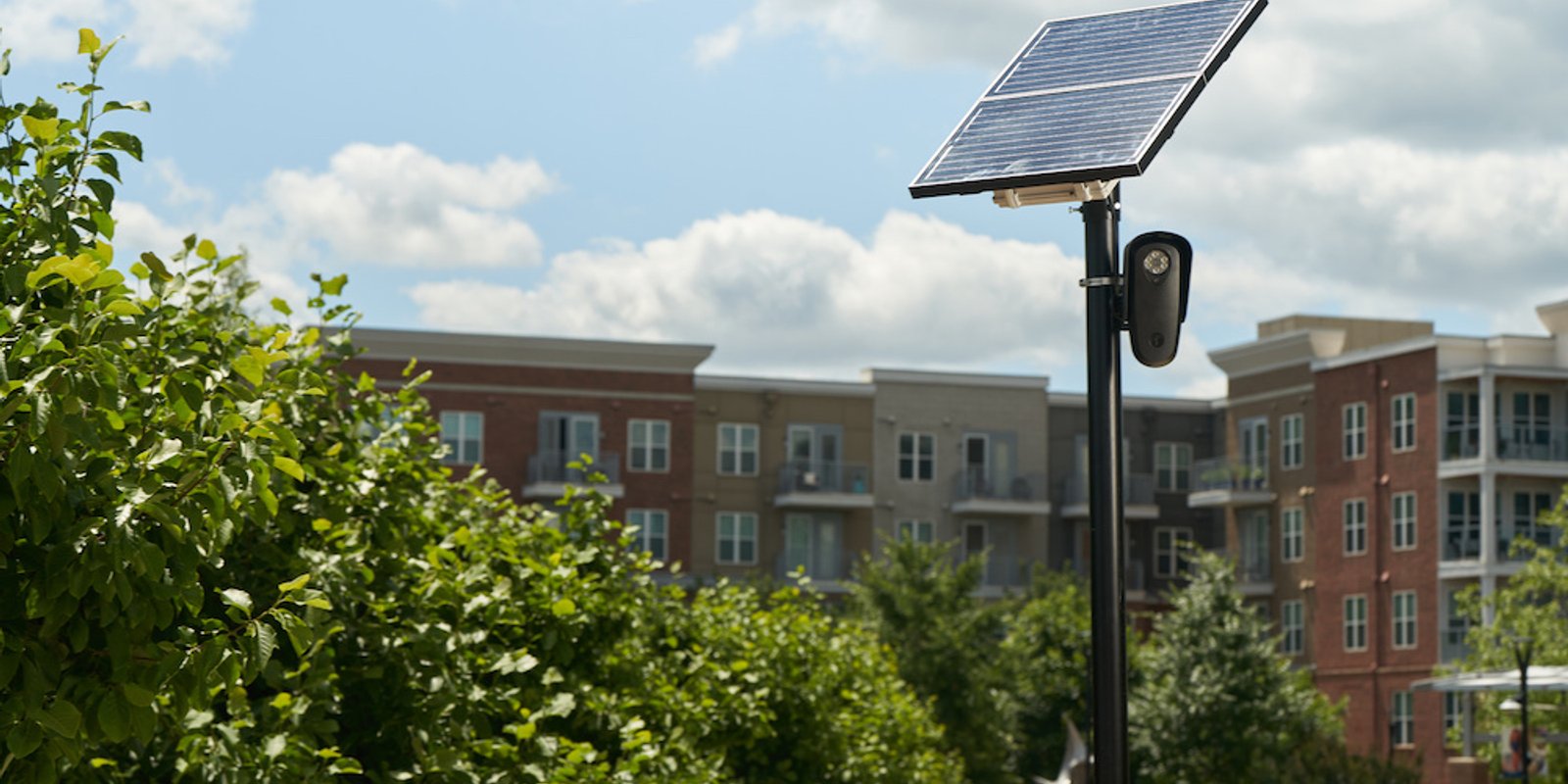 A solar-powered license plate reader camera on a pole in front of a brick apartment complex