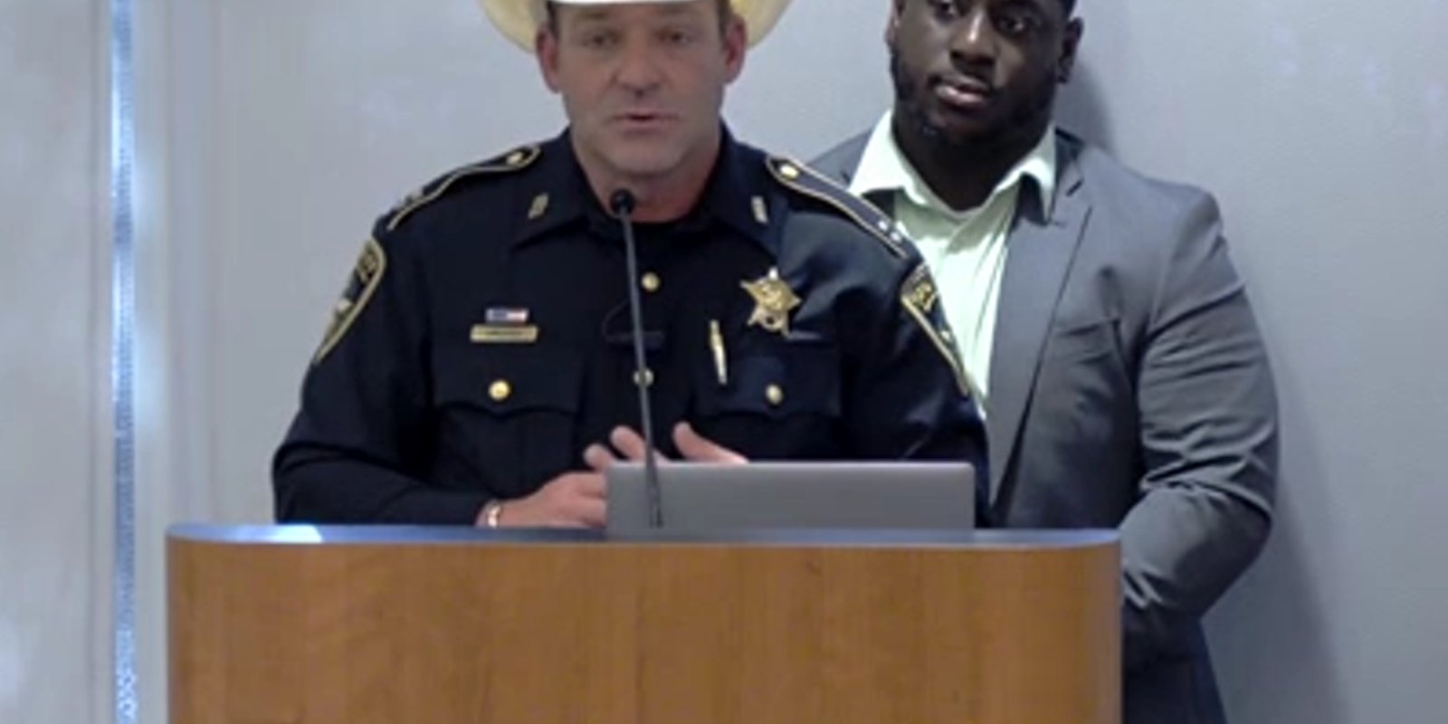 A sheriff’s deputy in a cowboy hat speaking at a podium microphone during a public meeting, with a man in a suit standing behind him