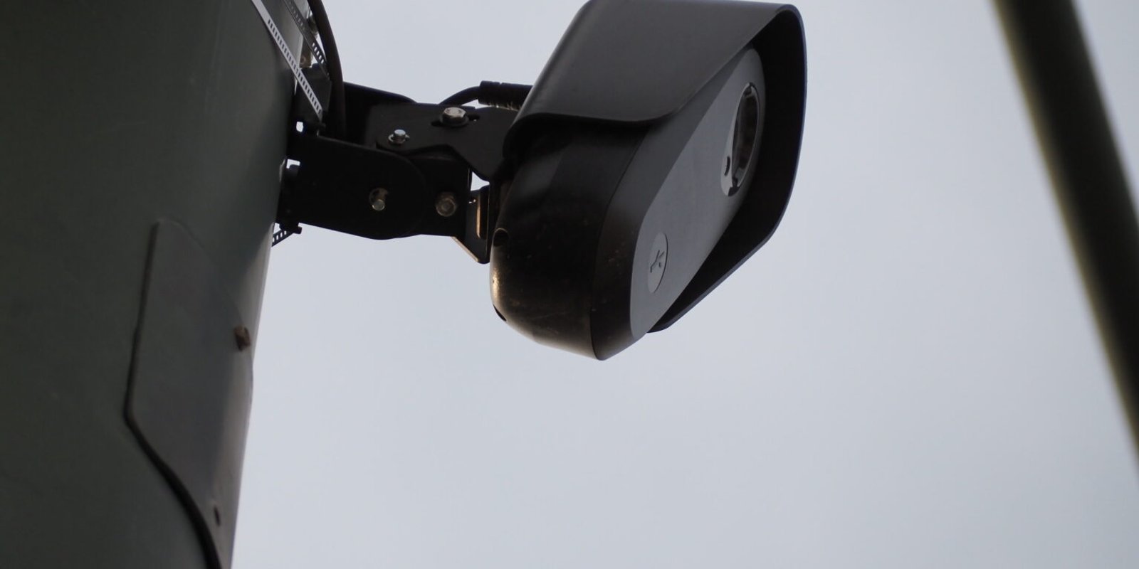 A close-up of a black automated license plate reader camera strapped to a utility pole against an overcast sky